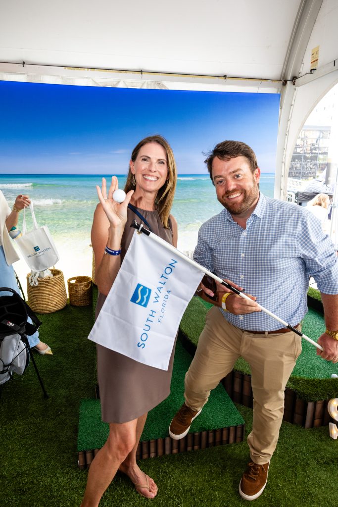 Smiling couple holding mini golf flag and ball inside event tent with ocean backdrop.
