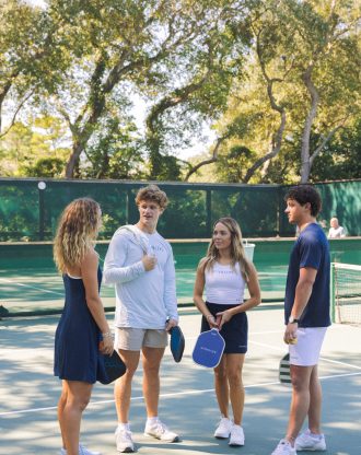 Four people standing on an outdoor pickleball court, talking while holding paddles.