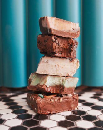 Stack of assorted chocolate fudge squares on black-and-white tile against turquoise wall.