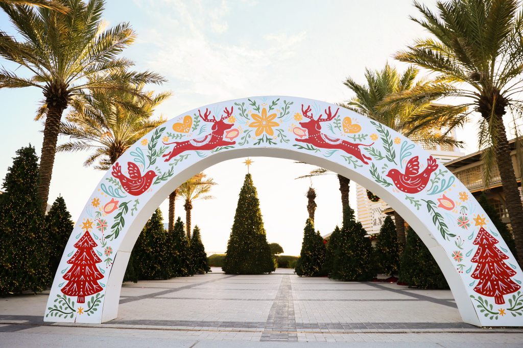 A festive holiday arch with red reindeer and floral designs frames palm trees and decorated Christmas trees outdoors.