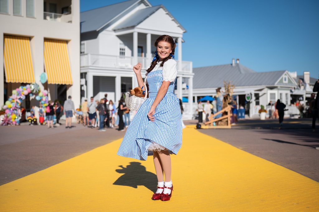 Woman in a blue gingham dress and red shoes poses on a yellow walkway at an outdoor village event with onlookers nearby.