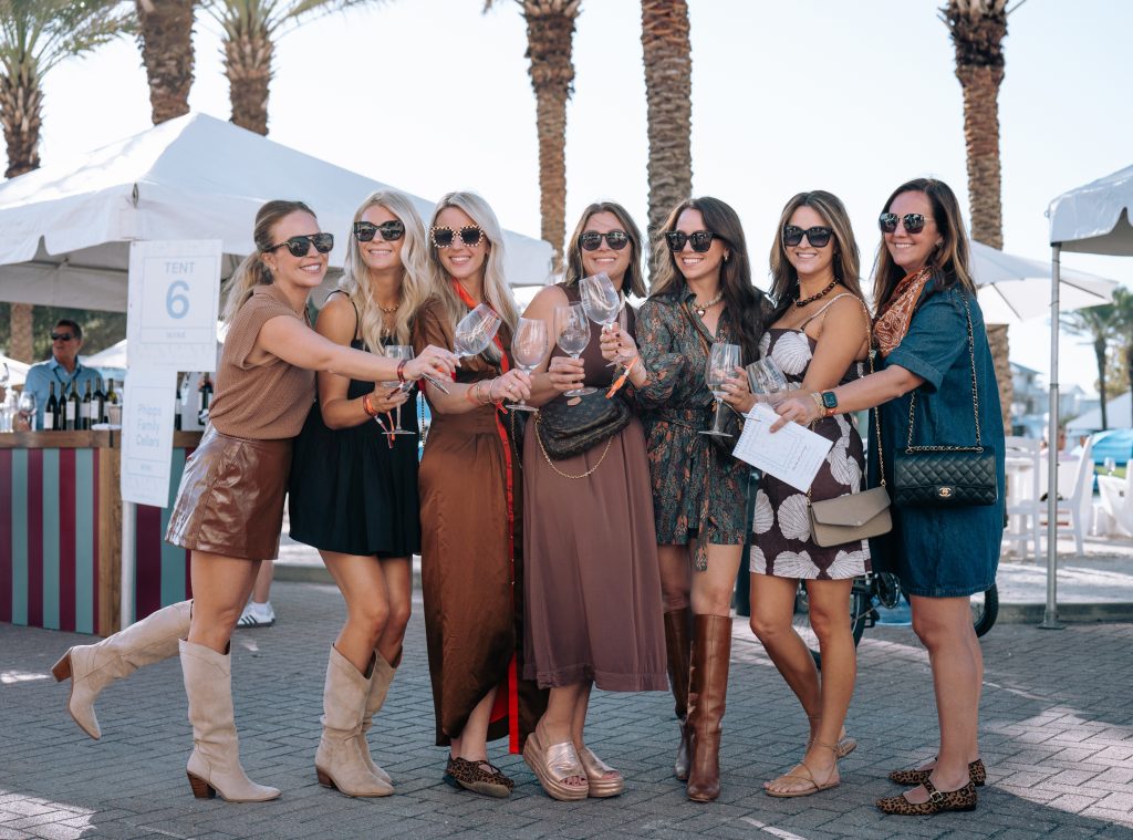 Group of women smiling and clinking wine glasses at an outdoor wine festival with palm trees and tents.