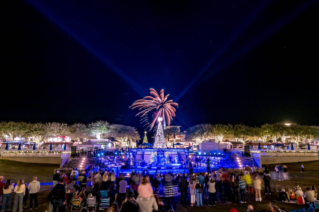 Fireworks burst above a nighttime outdoor concert as a crowd gathers around a lit stage and festive village lights.