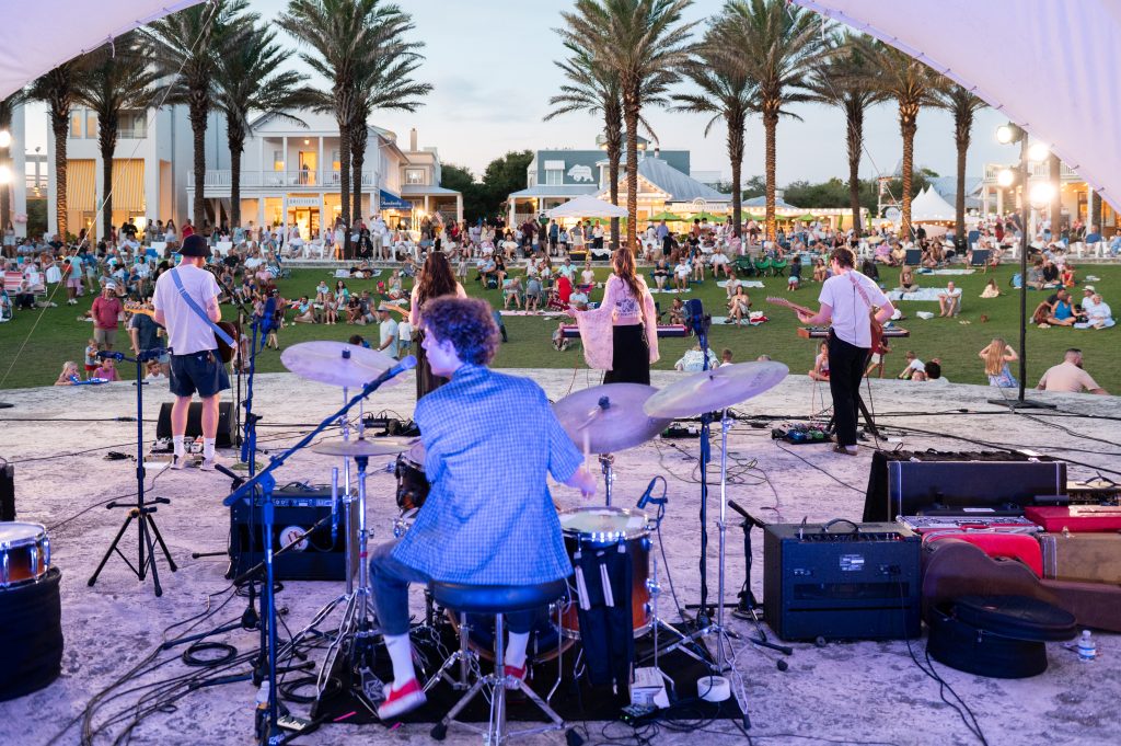 Live band performing on an outdoor stage facing a large crowd seated on grass beneath palm trees at sunset.