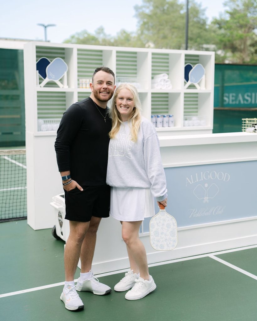 Two people smiling on a pickleball court holding paddles beside a light blue Allgood Pickleball Club stand