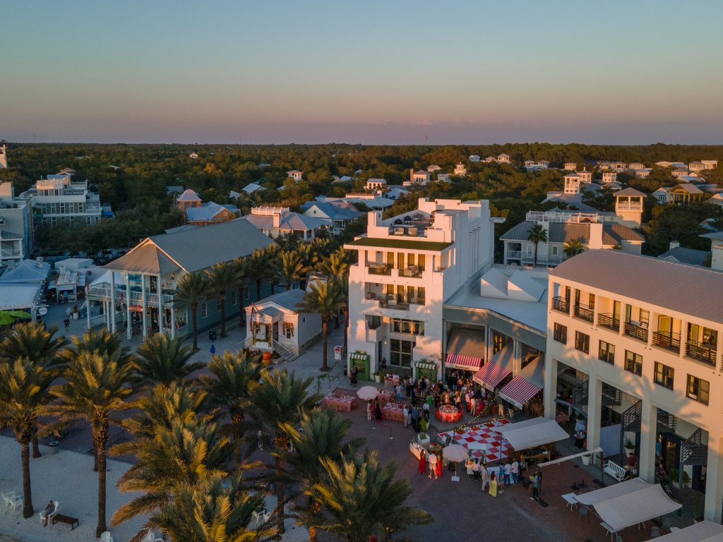 Aerial view of a coastal town at sunset with palm-lined streets, white buildings, and people gathered in a central plaza.