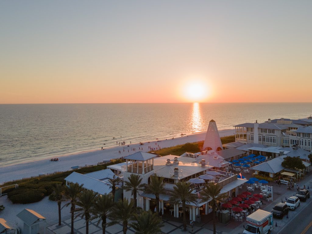 Sun setting over a beachfront resort with palm trees, white buildings, and the ocean stretching to the horizon.