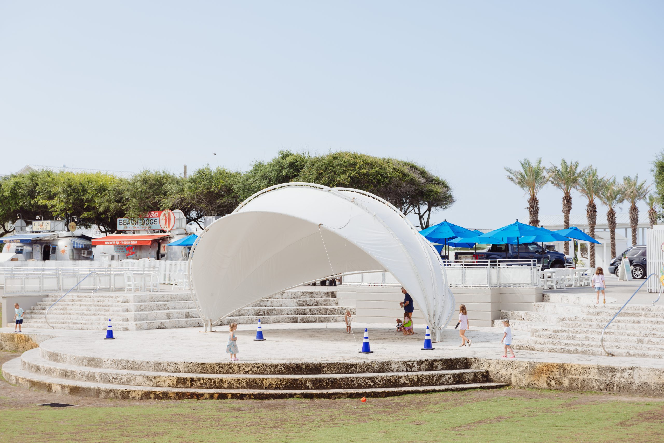 Children playing around a white canopy structure in an outdoor area with food trucks, umbrellas, and palm trees