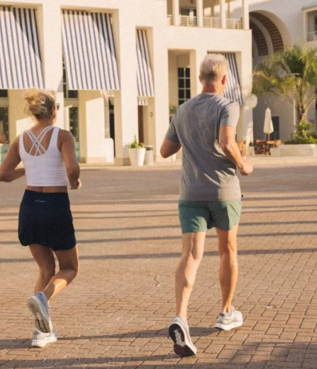 Man and woman jogging together on a sunny morning near modern white buildings with striped awnings.