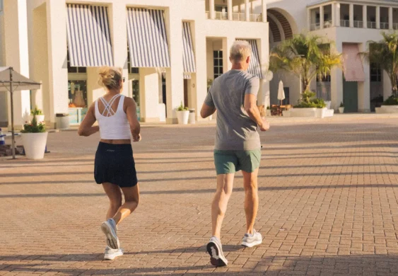 Man and woman jogging together on a sunny morning near modern white buildings with striped awnings.