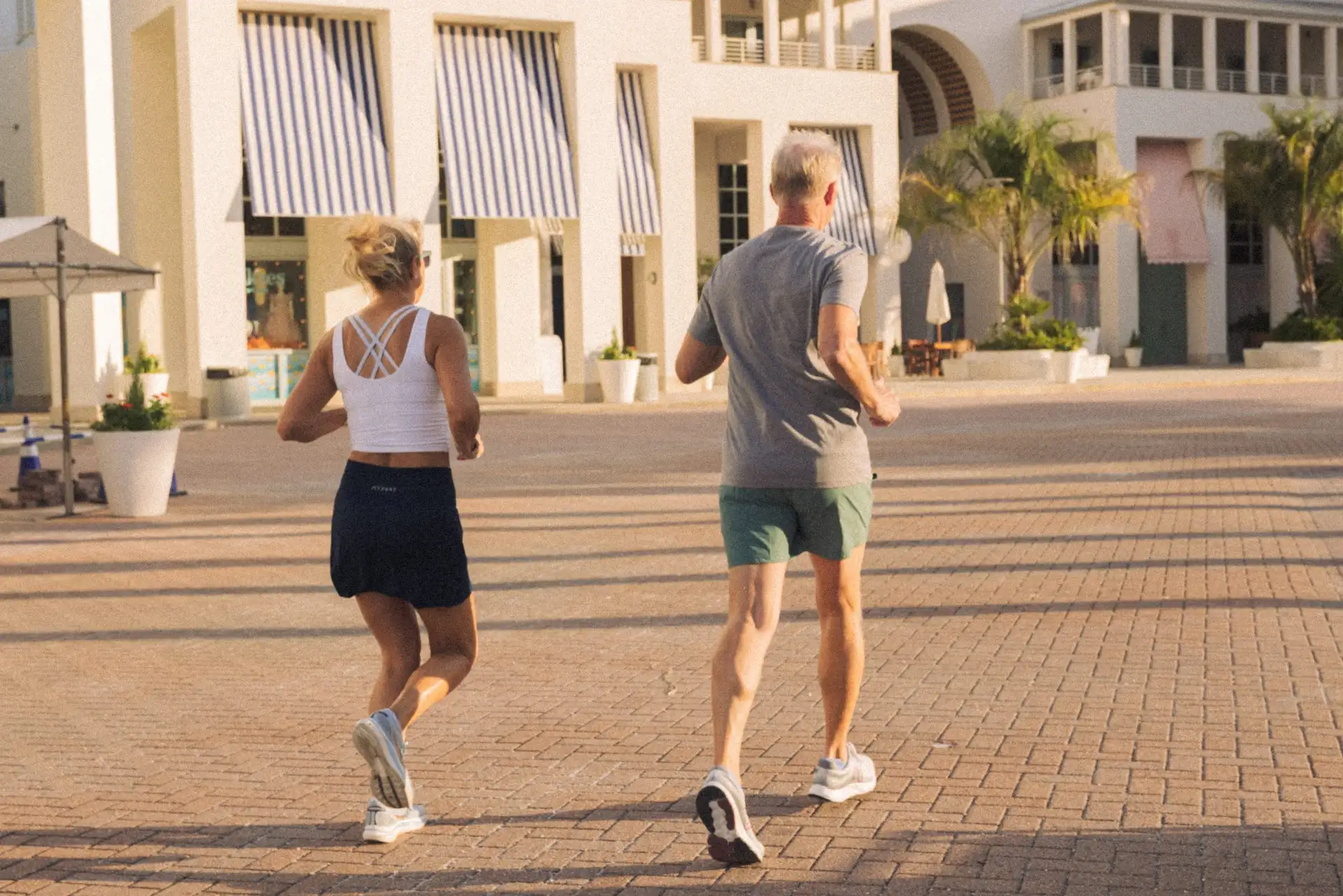 Man and woman jogging together on a sunny morning near modern white buildings with striped awnings.