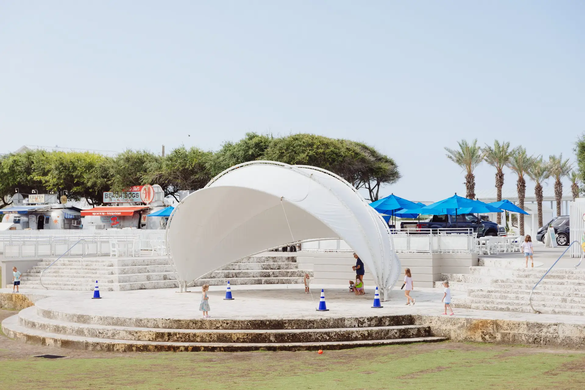 Children playing around a white canopy structure in an outdoor area with food trucks, umbrellas, and palm trees