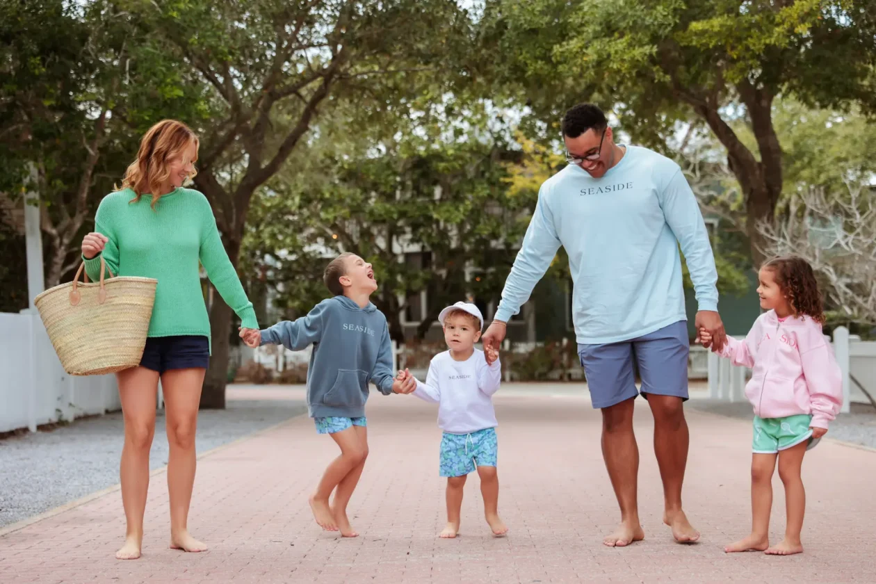 A happy family of five holding hands and walking barefoot down a brick pathway lined with trees.