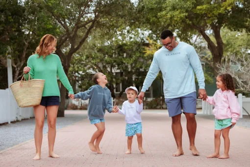 A happy family of five holding hands and walking barefoot down a brick pathway lined with trees.