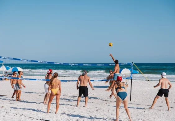 Group of people playing beach volleyball in swimwear on white sand, ocean and umbrellas in background.