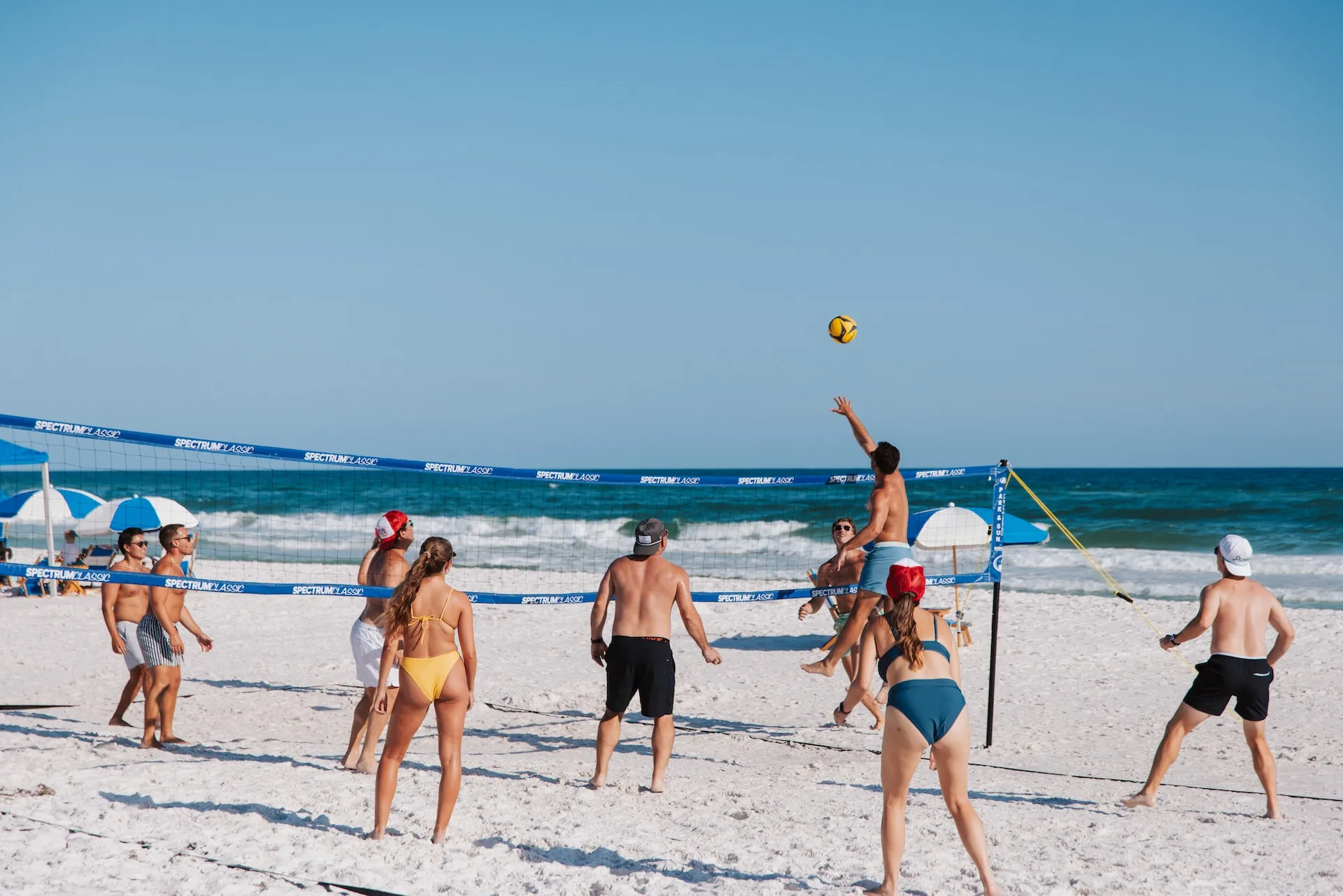 Group of people playing beach volleyball in swimwear on white sand, ocean and umbrellas in background.