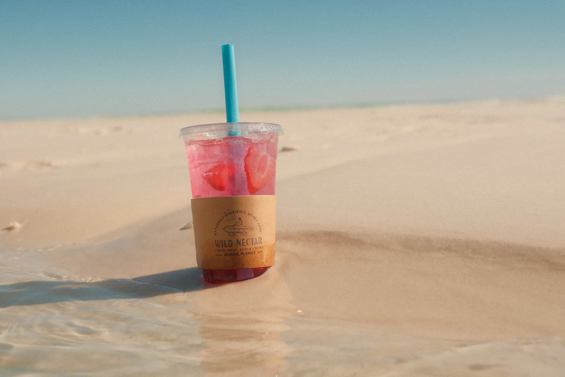 Pink iced drink with fruit slices in plastic cup resting on sunlit beach sand.