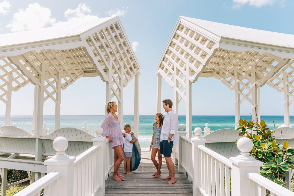Family of four standing on a white boardwalk pavilion overlooking a sunny turquoise beach and ocean.