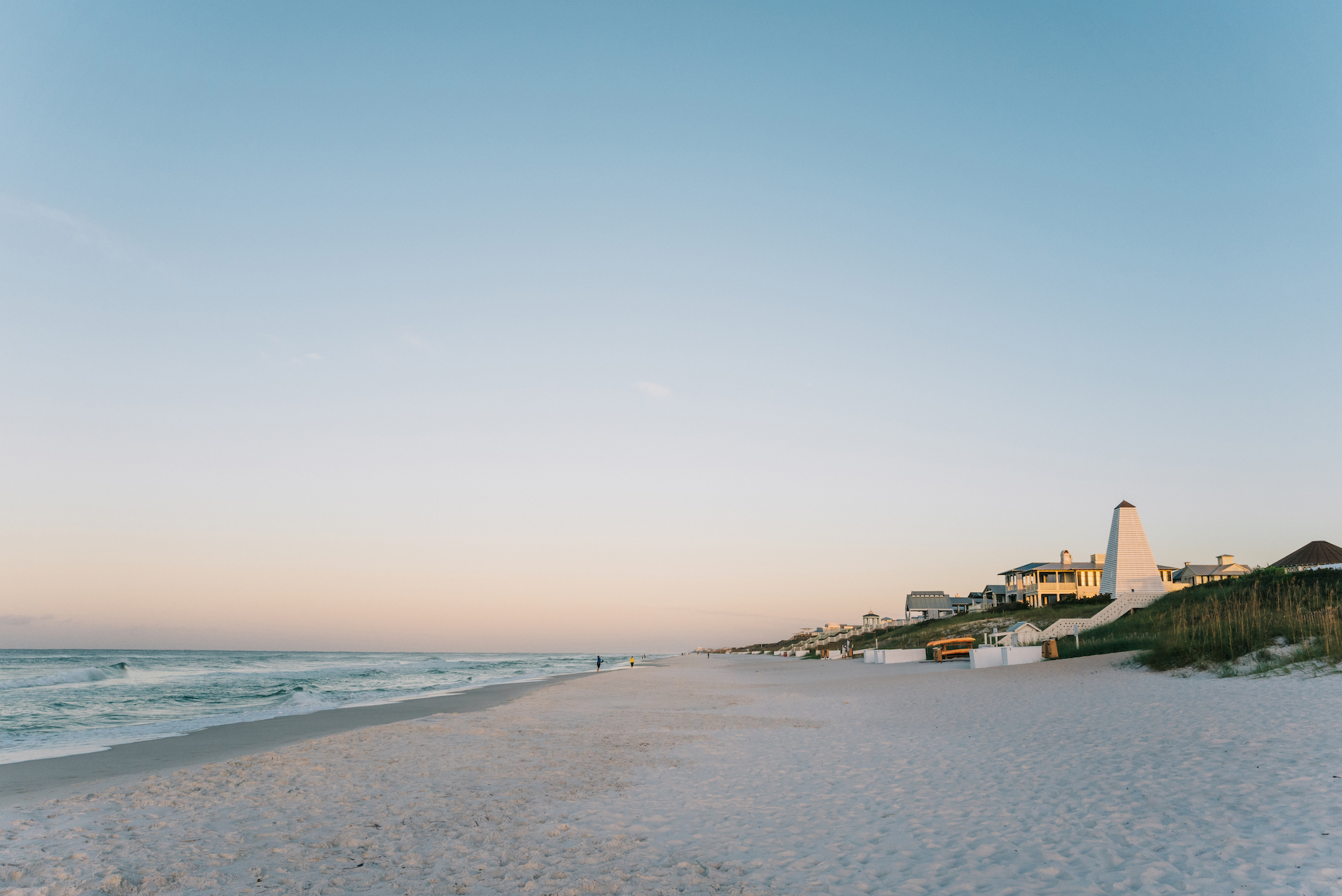Quiet white-sand beach at sunset with gentle waves, pastel sky, and coastal homes along the dunes.