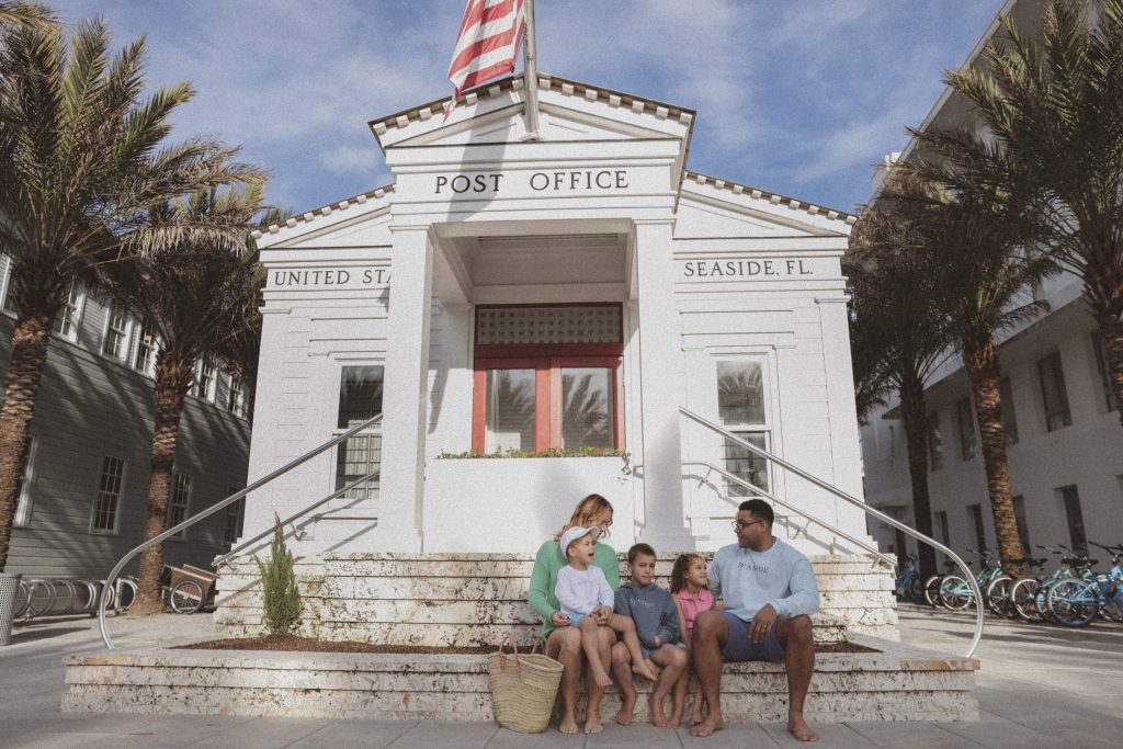 Family sitting on steps outside a white seaside post office lined with palm trees and bicycles.