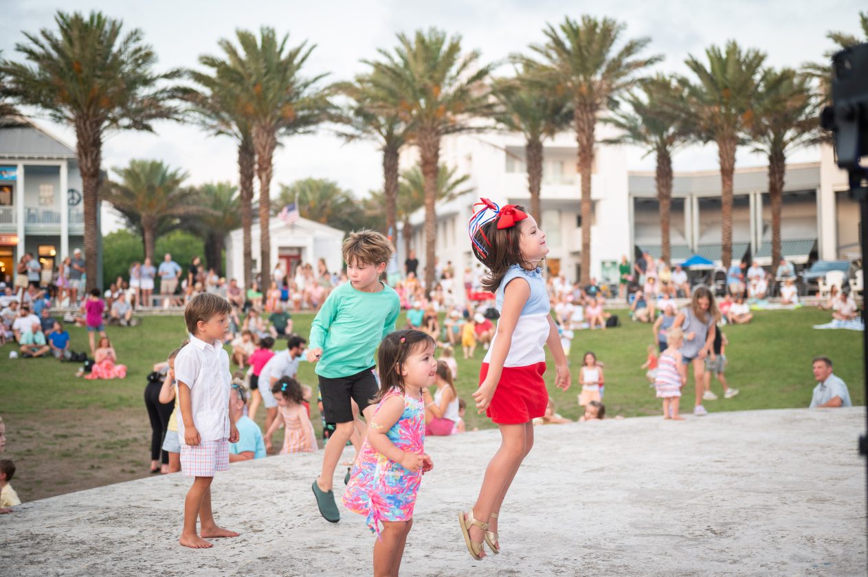 Children dance and play outdoors at a community gathering with palm trees and families in the background.