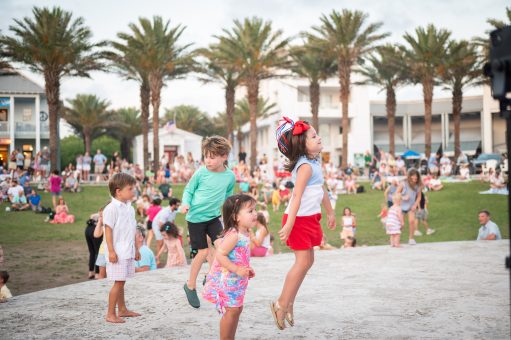 Children dance and play outdoors at a community gathering with palm trees and families in the background.