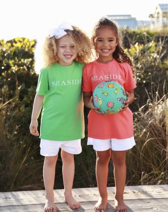 Two smiling children in colorful Seaside shirts stand barefoot outdoors, holding a patterned beach ball.