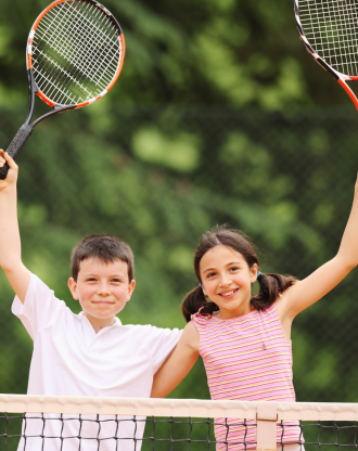 Smiling boy and girl standing at a tennis net, raising their rackets in celebration.