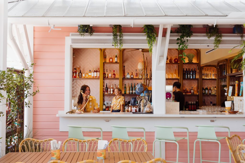 Claude responded: The tropical bar at Daytrader Tiki Bar & Restaurant in Seaside, Florida, featuring smiling staff behind a pink and gold-accented open-air bar stocked with spir…The tropical bar at Daytrader Tiki Bar & Restaurant in Seaside, Florida, featuring smiling staff behind a pink and gold-accented open-air bar stocked with spirits, surrounded by lush greenery and pastel outdoor seating on 30A.