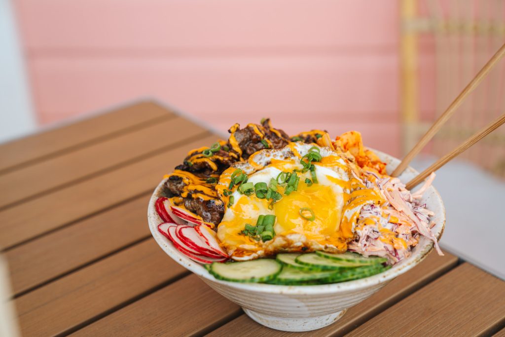 A Polynesian-inspired rice bowl at Daytrader Tiki Bar & Restaurant in Seaside, Florida, topped with a fried egg, grilled meat, spicy aioli drizzle, sliced radishes, cucumber, coleslaw, and green onions, served with chopsticks on an outdoor table along 30A.