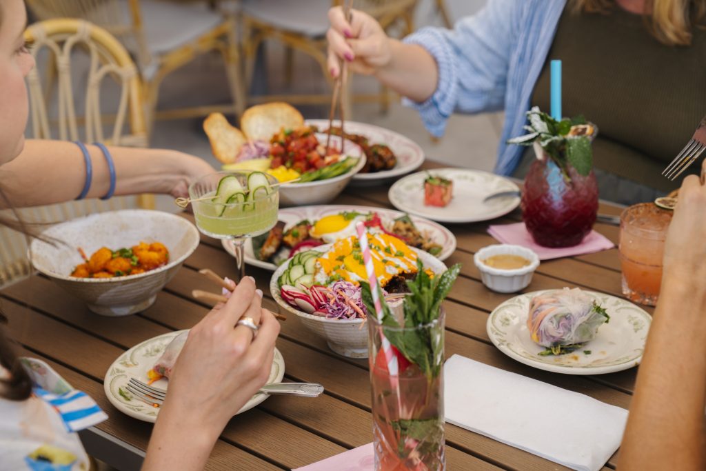 Friends dining together at Daytrader Tiki Bar & Restaurant in Seaside, Florida, sharing a spread of Polynesian-inspired rice bowls, fresh appetizers, and colorful tropical cocktails on the outdoor patio along 30A.