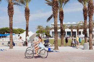 45 facts about seaside Woman in a light blue dress riding a bicycle down a palm tree-lined path near the beach, with beachgoers, umbrellas, and white coastal buildings in the background