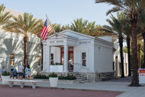 45 facts about seaside The white Seaside, Florida Post Office building with a columned porch and American flag, shaded by palm trees along a brick street.